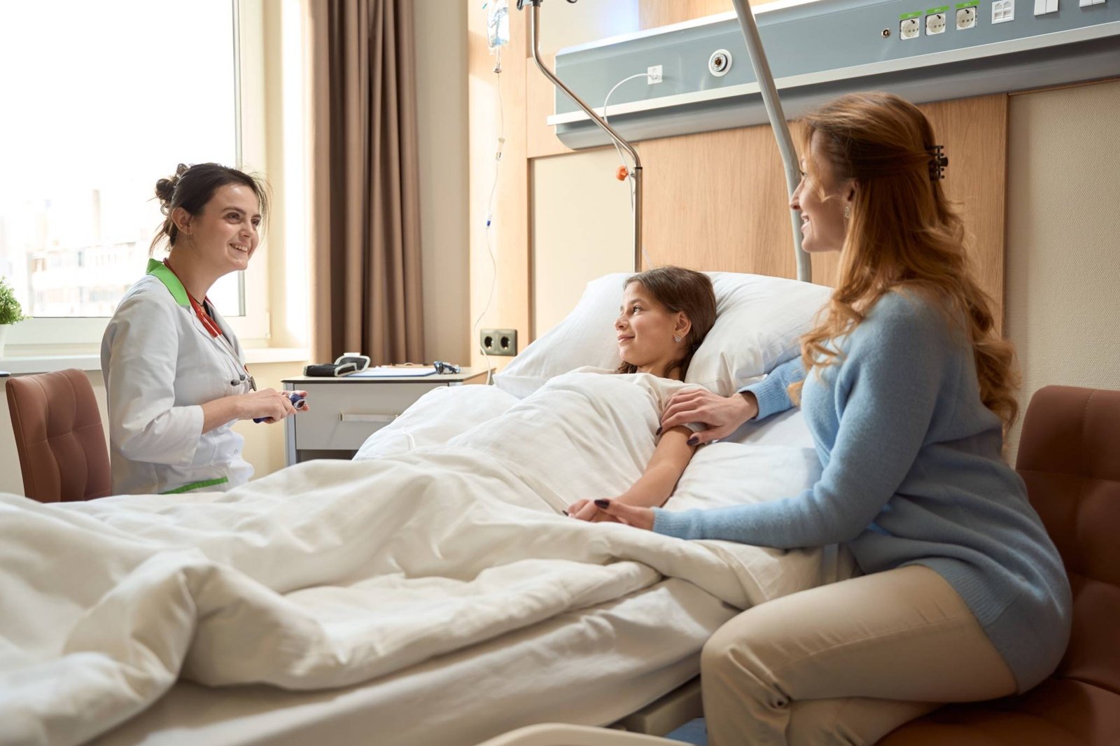 material-descanso-habitacion-infantil Paciente pediátrica en cama hospitalaria acompañada por su madre y atendida por una profesional sanitaria.
